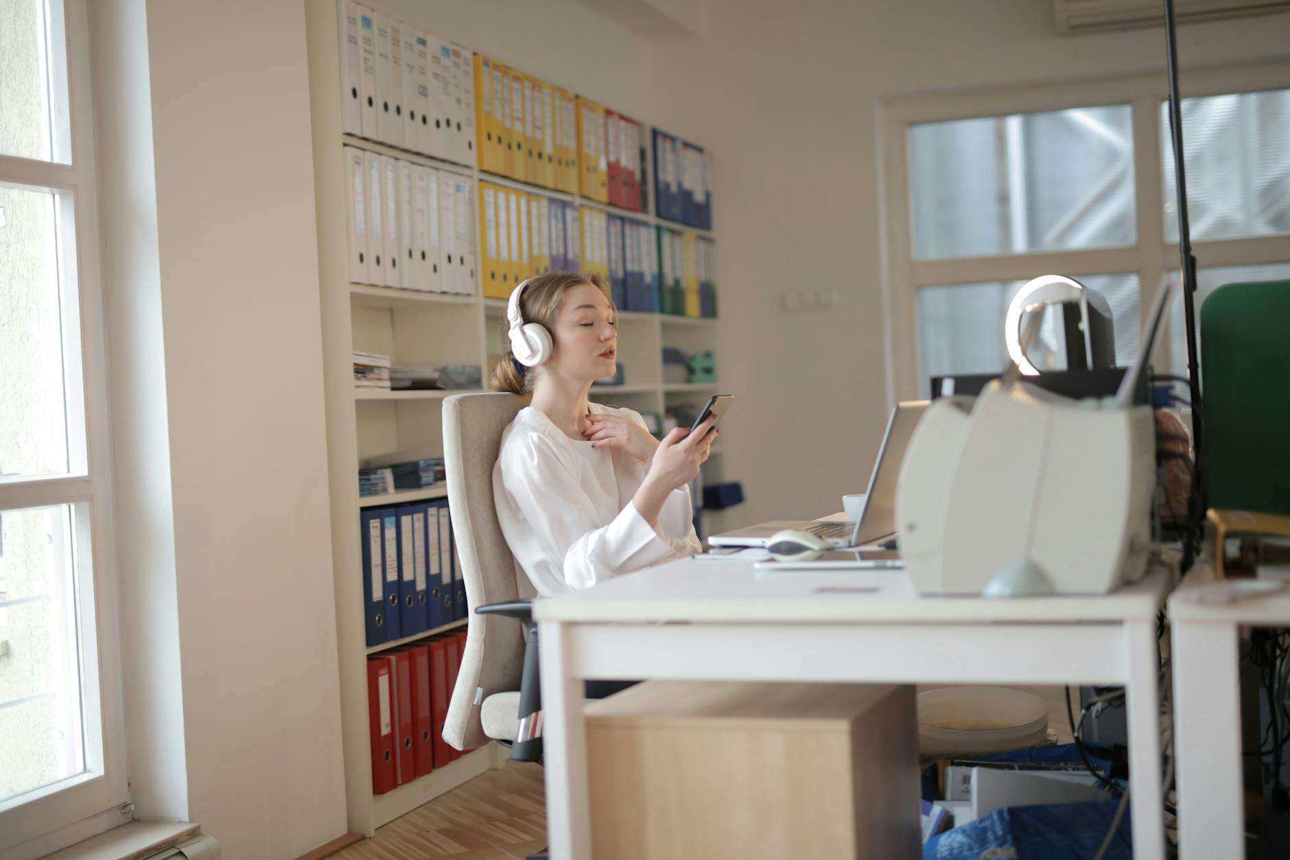 Woman using smartphone and headphones at modern office desk representing professional on-hold experience