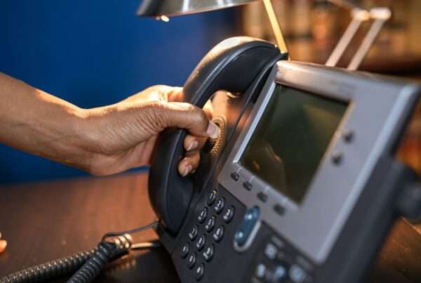 Close-up of a hand lifting the handset of an office desk phone, representing E911 emergency call testing and dispatchable location verification on a business phone system