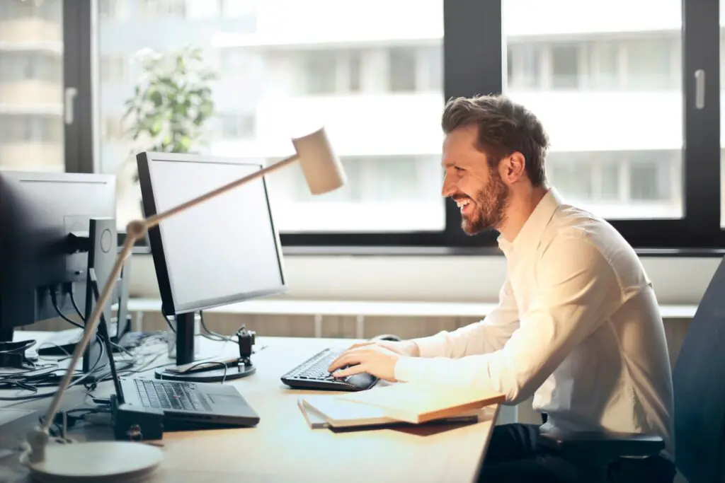 A business professional smiling while managing email at a desktop computer in a bright office.