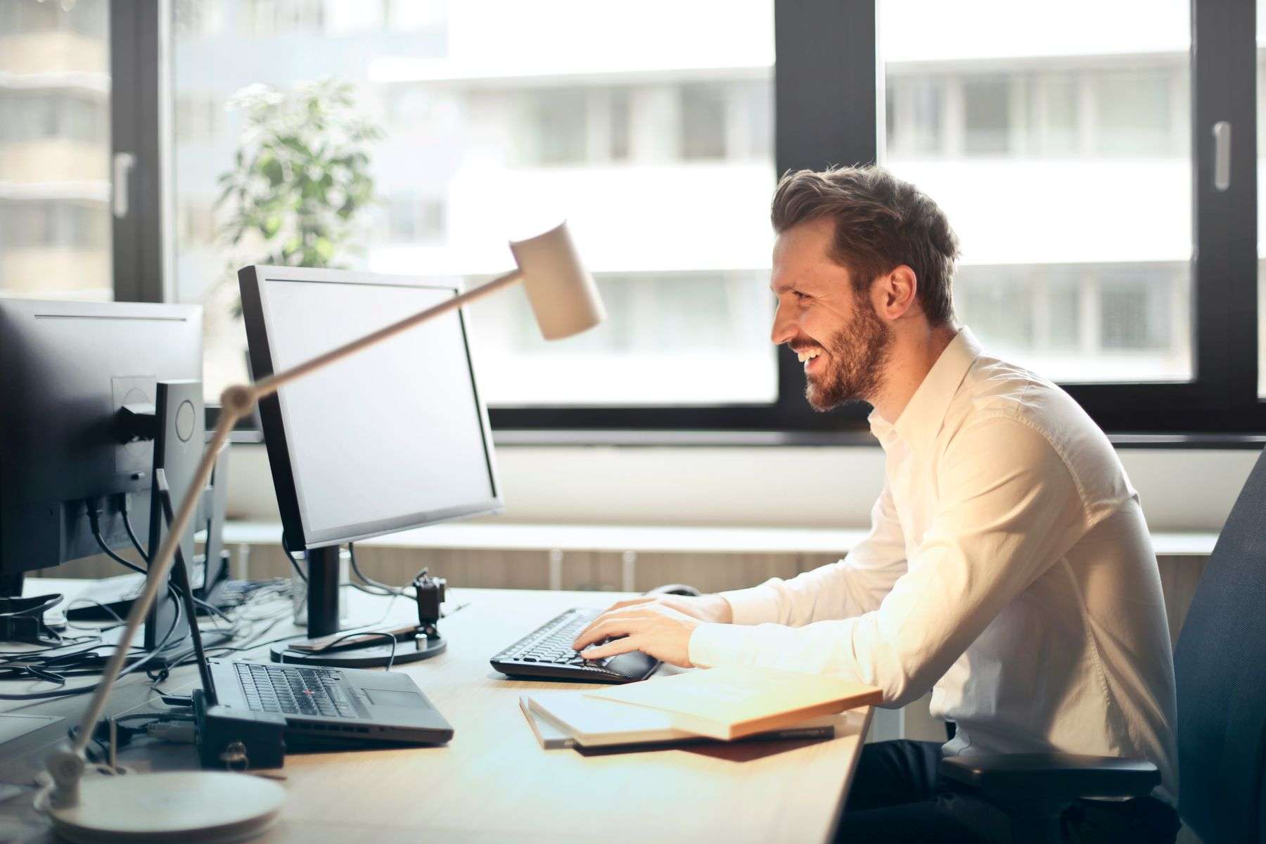 A business professional smiling while managing email at a desktop computer in a bright office.