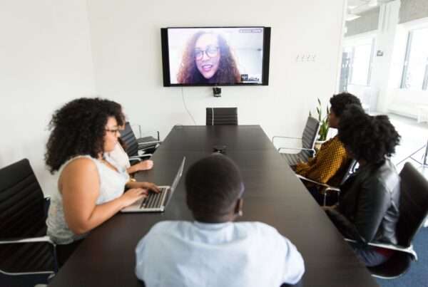 Coworkers gathered around a conference table collaborating with a remote teammate on a video call, illustrating hybrid team cloud communications