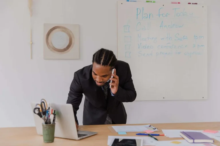 Businessman at an office desk answering a phone call while working on a laptop, representing a small business evaluating an answering service