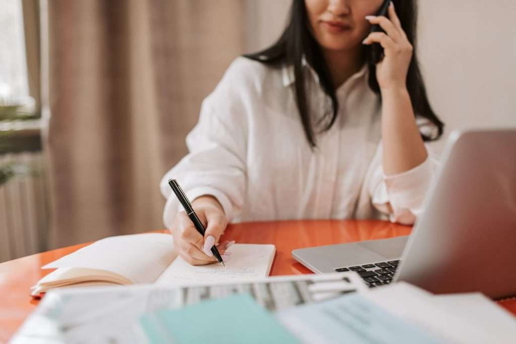 Professional in a white shirt writing notes in a notebook while talking on a smartphone beside a laptop
