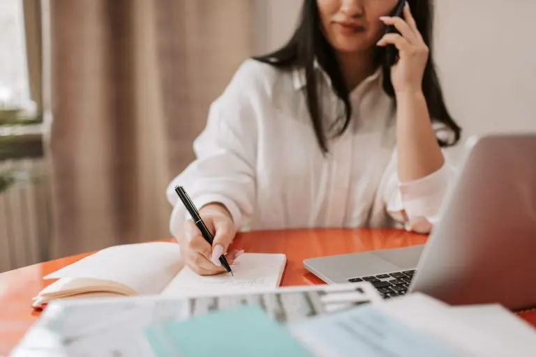 Professional in a white shirt writing notes in a notebook while talking on a smartphone beside a laptop