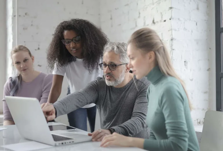 Four coworkers gathered around a laptop discussing meeting action items