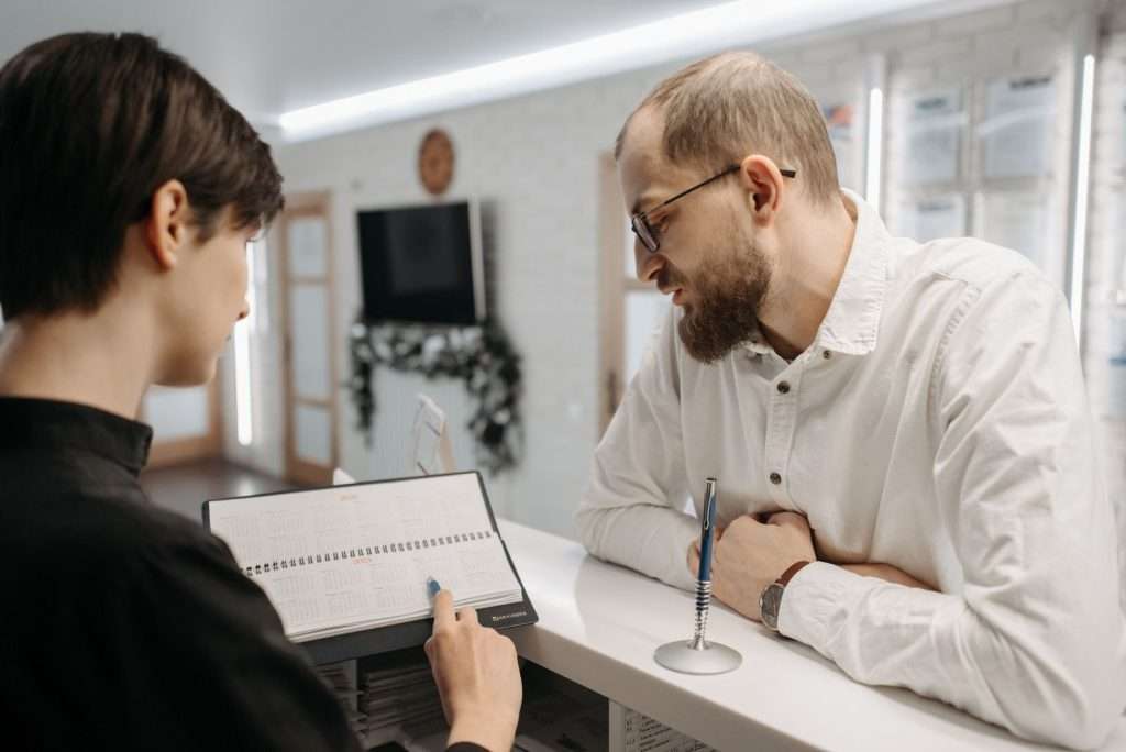 Medical clinic receptionist reviews an appointment book with a patient at a modern front desk